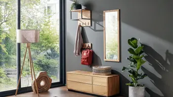 Garderobe aus Massivholz bestehend aus einem Spiegel, grifflose Schuhbank, Hutablage mit schwarzen Elementen, vor einer grauen Wand mit einer Pflanze und Stehlampe.