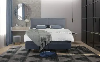 Modernes Boxspringbett in elegantem Blau, dekoriert mit passender Tagesdecke und Kissen, umgeben von einem geometrischen Teppich, Spiegelakzenten und minimalistischen Möbeln.
