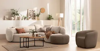 Creme farbenes rundes Ecksofa mit Anstellhocker in braun und rundem Sessel in braun in einem hellen gemütlichen Wohnzimmer mit mehreren beigen Vasen mit grünen Blumen und Grünpflanzen.