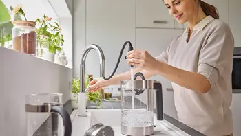 Junge Frau füllt einen Wasserkocher an einer modernen Küchenarmatur mit ausziehbarem Schlauch in heller, freundlicher Küche mit Pflanzen auf der Fensterbank.