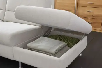 Rechter Teil eines beigen Sofas, bei dem die Sitzfläche nach oben geklappt ist und den Blick auf einen Stauraum in dem Sofa freigibt, dort sind Decken gelagert.