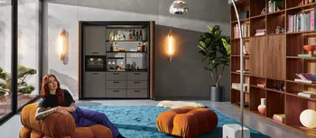 Frau auf Sitzkissen in Wohnung mit kompakter Kuecheneinheit von Next125 vor Buecherregal.