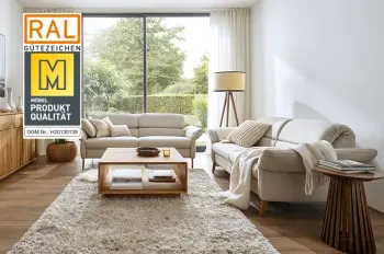 Gütesiegel "Goldene M" auf einem Bild von zwei beigen Sofas mit hellen Kissen und Decke in einem hellen Wohnzimmer mit großer Pflanze, Stehlampe und Couchtisch.