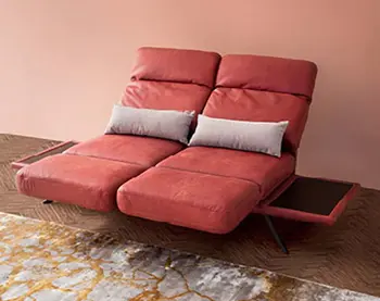 Modernes rotes Zweisitzer-Sofa mit Relaxfunktion, zwei grauen Kissen und seitlichen Ablageflächen vor rosafarbener Wand und gemustertem Teppich