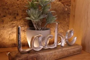Dekoratives Schild mit dem Schriftzug „lache“ aus Metall auf einem Holzsockel, dahinter eine kleine grüne Pflanze in einem grauen Topf.