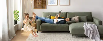 Helles Wohnzimmer mit einem grünen Ecksofa, auf dem ein Mann gemütlich liegt und lächelnd auf seine beiden Kinder im Raum sieht.