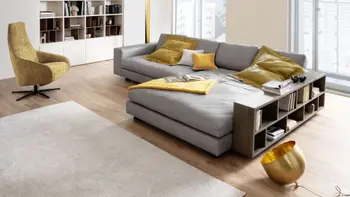 Graues modernes Ecksofa mit Lederbezug, auf dem gelbe und ein weißes Kissen und eine gelbe Decke liegen und neben dem ein Regal steht.