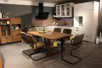 Gemütlicher Essbereich mit Massivholztisch und sechs gepolsterten Stühlen vor einer modernen Einbauküche in Weiß und Holzoptik.