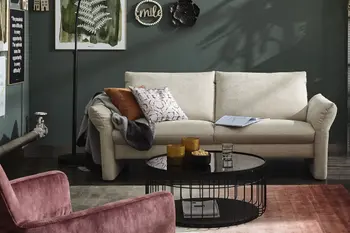 Modernes beiges Sofa, auf dem zwei Kissen, ein Buch und eine Decke liegen vor einer grünen Wand.