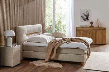 Boxspringbett mit beigem Stoffbezug daneben ein Nachttischkästchen mit dem gleichen Stoffbezug vor einer Wand mit Akkustikpaneelen