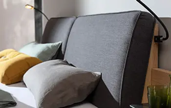 Modernes Bett mit gepolstertem, grauem Kopfteil und integrierter Lesebeleuchtung – perfekter Komfort für entspannte Leseabende im Schlafzimmer mit stilvollen Kissen in Gelb, Grau und Grün.
