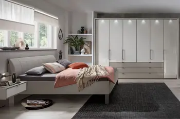 Heller Raum mit weißem Bett und passenden Nachtkonsolen und großem weißen Kleiderschrank mit Türen und Schubladen und Beleuchtung oberhalb der Türen.