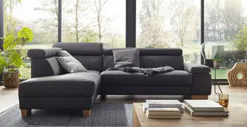 Graues Ecksofa aus Leder mit klappbaren Armlehnen und Kopfstützen dekoriert mit grauen Kissen und einem grauen Fell vor großen Fenstern.