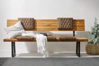 Sitzbank aus Massivholz mit Kufen aus Metall, braune gesteppte Rückenkissen an der Rückenlehne und auf der Bank sowie einer Decke und Kissen in hellgrau, daneben steht eine Pflanze.