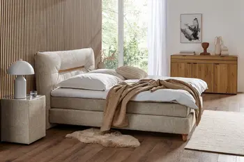 Boxspringbett in beigem Stoff mit Kissen in beige und einer weißen Decke und einer camelfarbenen vor einer Wand mit Akkustikpaneelen.