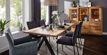 Massivholz Esstisch mit Gestell aus schwarzem Metall, um den herum Stühle in grautönen mit und ohne Armlehne stehen in einem gemütlichen Esszimmer mit beigem Teppich und Massivholz Highboard.