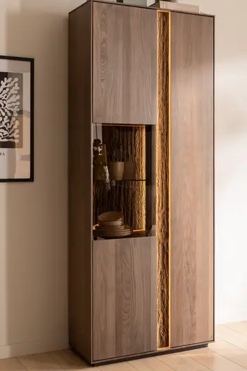 Moderne hohe Vitrine aus massiver Buche mit zwei Türen, in der linken Türe befindet sich ein Glaselement, das beleuchtet ist.