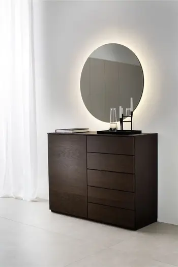 Runder beleuchteter Spiegel unter dem eine braune Designer Kommode steht, die links eine Türe hat und auf der rechten Seite fünf Schubladen.