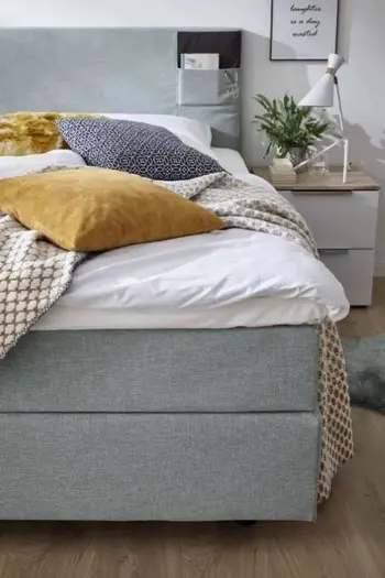 Rechter Teil eines grauen Boxspringbettes mit einer Aufbewahrungstasch eim Kopfteil, dekoriert mit weißer Decke und gelben und blau-gemustertem Kissen und gelb weißen Wolldecke.