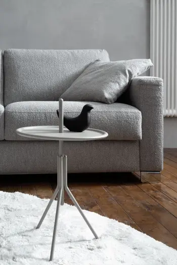 Rechte Ecke eines grauen Sofas mit großen grauen Rückenkissen und einem grauen Dekokissen, davor steht ein runder kleiner Bestelltisch mit einem schwarzen Dekovogel.