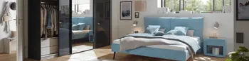 Hellblaues Doppelbett in einem hellen großräumigen Schlafzimmer mit verglastem Kleiderschrank und zum Bett passenden Beistelltischen.