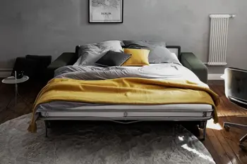 Grünes Schlafsofa, das zum Bett umgebaut ist mit einer großen Liegefläche mit Matratze, auf dem Decken und Kissen in gelb, grau, grün und weiß liegen.