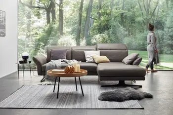 Graues Ecksofa aus Leder mit klappbaren Kopfstützen vor einer großen Fensterfront mit Blick auf Bäume.