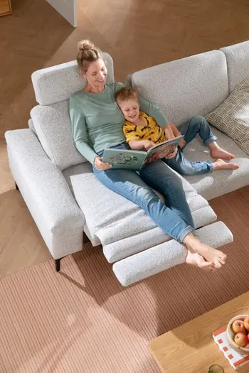 Eine Frau liegt mit einem Kleinkind auf einem Sofa mit Relaxfunktion und liest ein Buch vor.