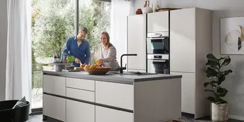 Mann und Frau in grauer Inselkueche mit Einbaugeraeten und Schneidebrett und Obst.