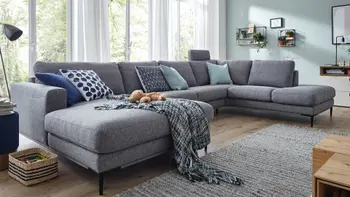 Graue Wohnlandschaft mit einer Kopfstütze in einem hellen Raum mit großen Fenstern.