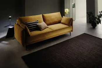Cognac farbenes modernes Zweisitzer Sofa in einem dunklen Raum, neben dem Sofa steht ein schwarzer Beistelltisch mit einer kleinen Tischlampe, die angeschalten ist.