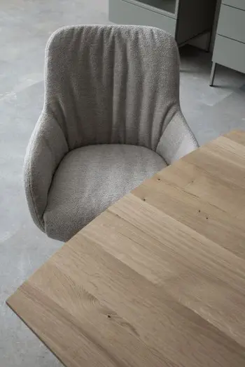 Moderner Esszimmerstuhl mit Armlehnen und beigem Stoff bezogen und moderner Faltenbildung an der Rückenlehne, der seitlich eines Esstisches steht.