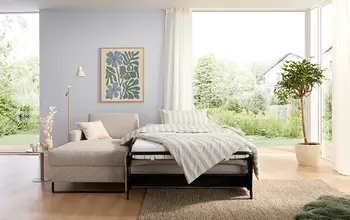 Helles Wohnzimmer mit ausziehbarem Schlafsofa in Beige vor bodentiefem Fenster mit Blick ins Grüne – moderne, platzsparende Schlafgelegenheit mit stilvoller Einrichtung.