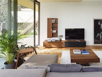 Helles Wohnzimmer mit großer Fensterfront mit Sicht auf einen Garten. Im Wohnzimmer an einer weißen Wand, ein Wandregal und ein Sideboard aus dunklem Holz. Davor ein TV-Schrank im passenden Look.