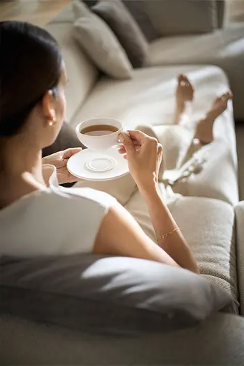 Frau liegt auf beigem Sofa und trinkt entspannt eine Tasse Kaffee.