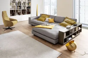 Graues modernes Ecksofa mit Lederbezug, auf dem gelbe und ein weißes Kissen und eine gelbe Decke liegen und neben dem ein Regal steht.
