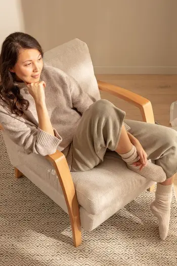 Frau sitzt auf einem beigen Stuhl mit Holzarmlehnen und blickt in die Ferne.