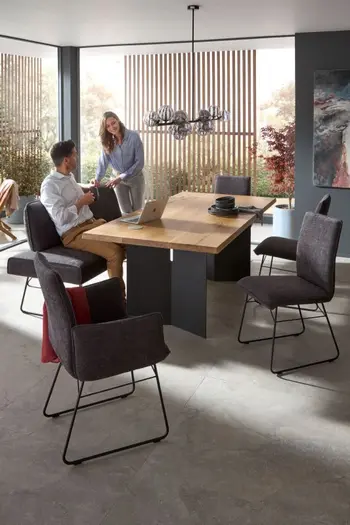 Modernes Esszimmer mit grauer Sitzbank und grauen Stühlen mit und ohne Armlehne und schwarzem Metallgestell um einen Massivholz Esstisch, auf der Bank sitzt ein Mann und eine Frau steht daneben.