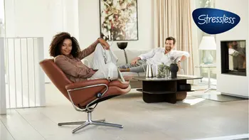 Frau sitzt entspannt in einem Stressless Rome Sessel in der Farbe Paloma Dark Henna, während ein Mann auf einem modernen Sofa im Hintergrund sitzt. Das Wohnzimmer ist hell und modern eingerichtet.