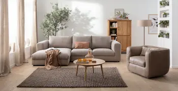 Graues Ecksofa mit großen grauen Kissen als Rückenpolster, dekoriert mit einer Decke und Kissen in rose, daneben ein passender Sessel in braunem Leder.