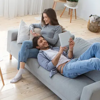 Ein glückliches Paar sitzt gemütlich auf einem Sofa im Wohnzimmer und surft im Internet mit Handy und Tablet.&nbsp;