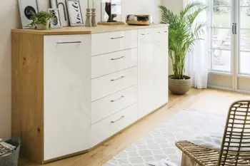 Große Kommode aus Massivholz mit einer Abschrägung links und Fronten in weiß Hochglanz und silbernen Griffen, dekoriert mit Bildern, Kerzenständern und einer Pflanze in einem hellen Raum.