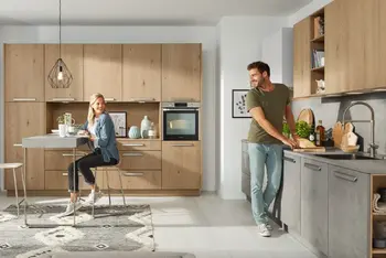 Ein Paar in der Küche schaut sich glücklich an. Die Frau sitzt an einem Tisch, der in das Schranksystem der Küche integriert ist. Der Mann steht an einer Küchenzeile und kocht.&nbsp;