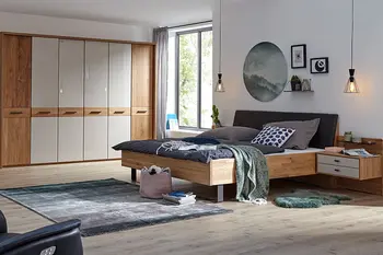 Holzbett mit daran befestigter Nachtkonsole, die weiße Schubladenfronten hat neben einem großen Kleiderschrank aus hellem Holz mit weißen Akzenten an den Türen.