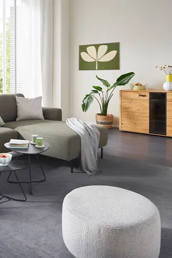 Beiger Hocker in einem hellen Wohnzimmer mit grünem Ecksofa. 