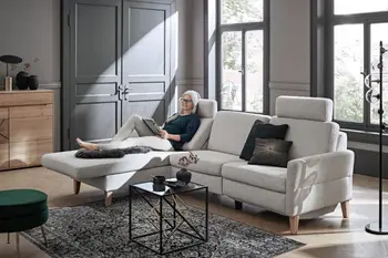 Graues Ecksofa mit Stoffbezug, auf dem eine ältere Frau liegt mit klappbaren Kopfstützen in einem Wohnzimmer mit großen Fenstern.