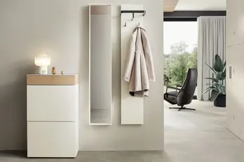 Weiße moderne Design Garderobenmöbel bestehend aus Highboard mit Holzelement, schmalem hängenden Spiegelschrank und hängendem weißen Brett mit schwarzen Garderobenhaken an dem ein Trenchcoat hängt.