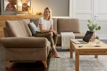 Hellbraunes Ecksofa mit Holzbeinen, auf dem eine Frau sitzt vor einem Laptop auf dem Couchtisch aus Massivholz.