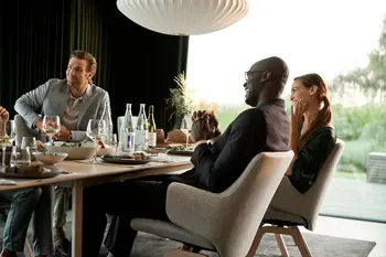 Eine Gruppe von Menschen sitzt auf Stressless Laurel Esszimmerstühlen in Clover Light Beige Stoffbezug, während sie gemeinsam an einem gedeckten Esstisch in einem eleganten Raum mit großer Fensterfront speisen und lachen.