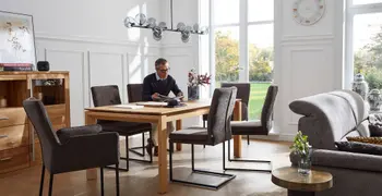 Mehrere graue Freischwinger mit schwarzem Metallgestell und zwei graue Armlehnstühle um einen Holz Esstisch, auf einem der Stühle sitzt ein Mann und liest.